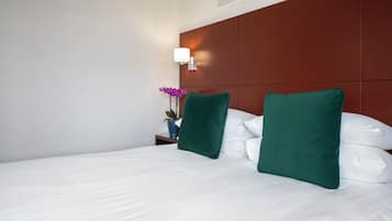Una cama bien tendida con sábanas blancas y dos almohadas verdes, una mesita de noche con una planta en maceta, y una lámpara de pared.