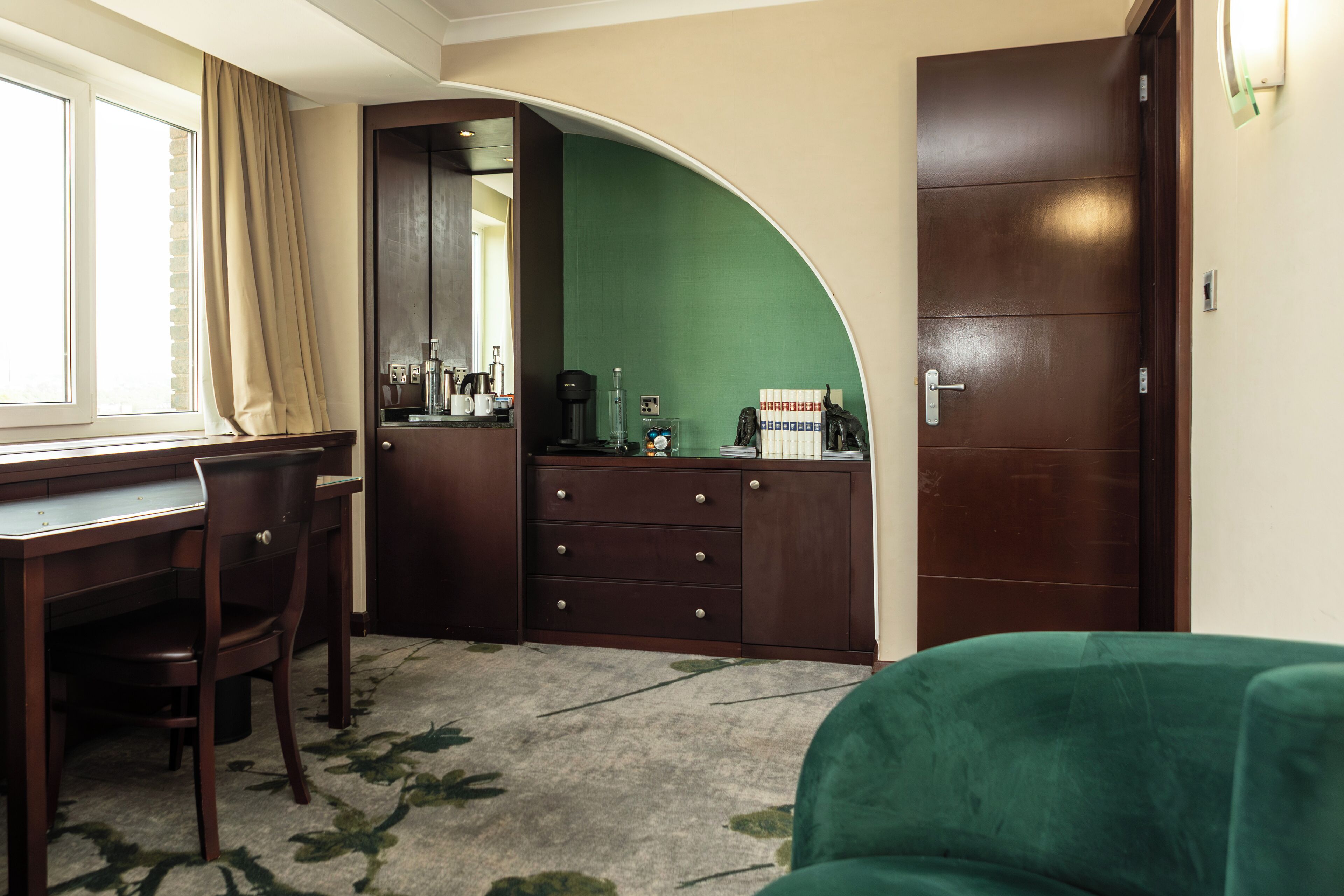 Une pièce comprenant un bureau, une chaise et un canapé vert. On y trouve également une armoire avec un miroir et un mur vert.