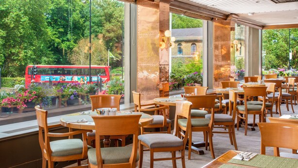 A restaurant with wooden tables and chairs, large windows with a view of a red bus and greenery outside. - Danubius Hotel Regents Park (London)