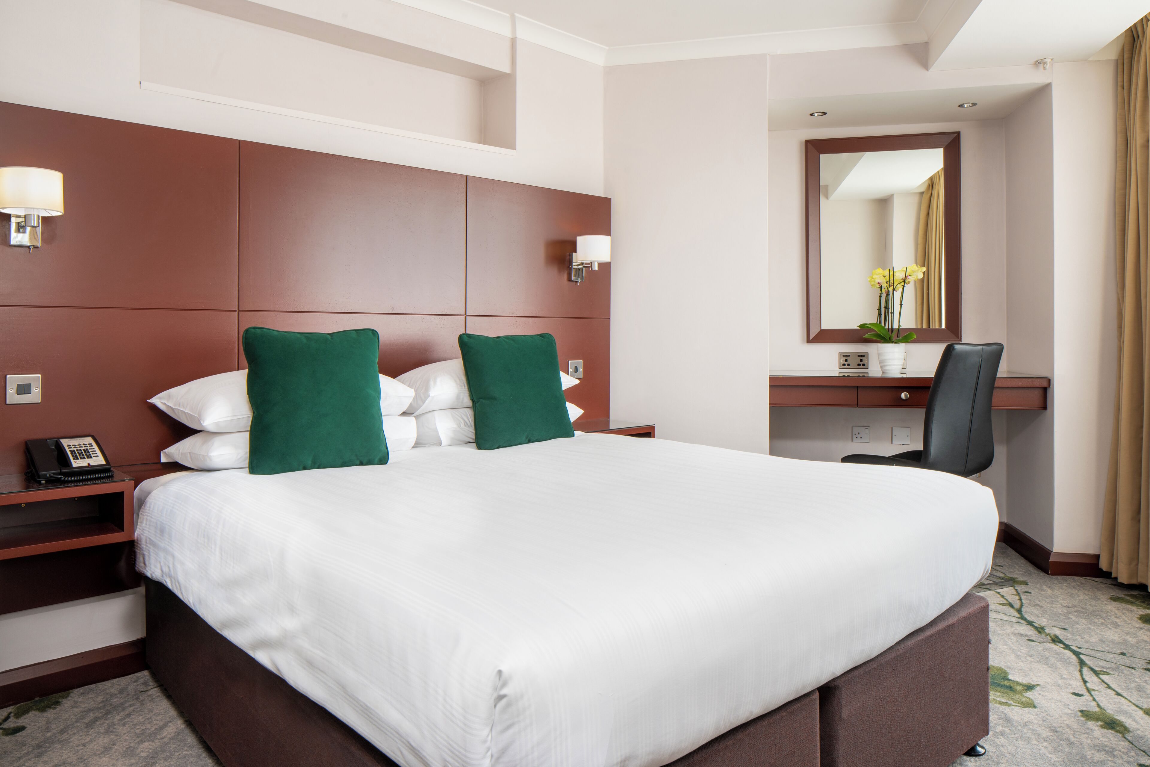 Quarto de hotel com uma cama grande, duas almofadas verdes, uma escrivaninha com cadeira, um espelho e telefone na mesinha de cabeceira.