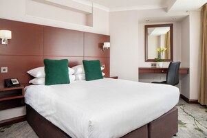 A hotel room with a large bed, two green pillows, a desk with a chair, a mirror, and a telephone on the side table.