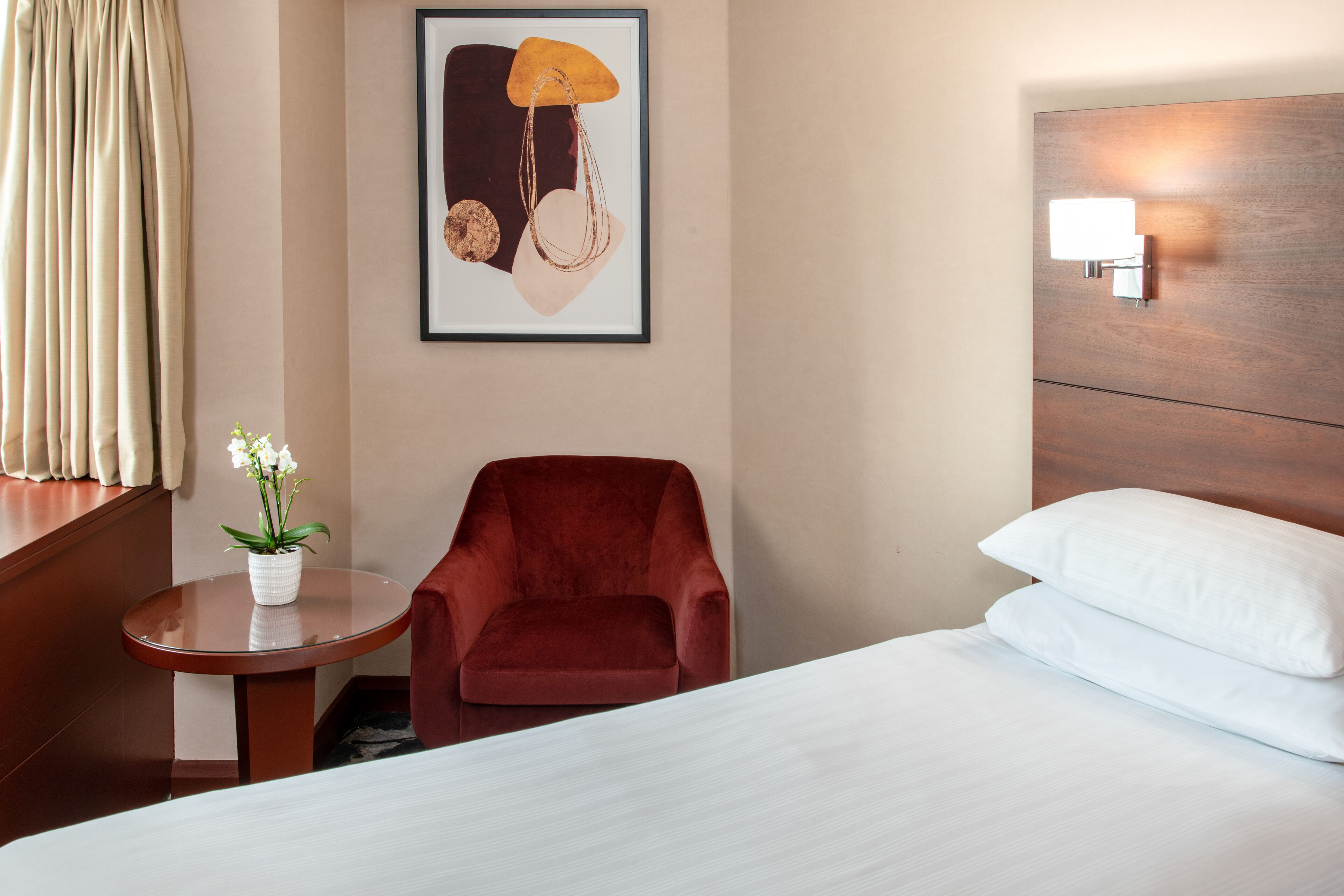 Una habitación de hotel con una cama, un sillón rojo, una mesita con un jarrón de flores, una obra de arte enmarcada en la pared y una mesita de noche con una lámpara.