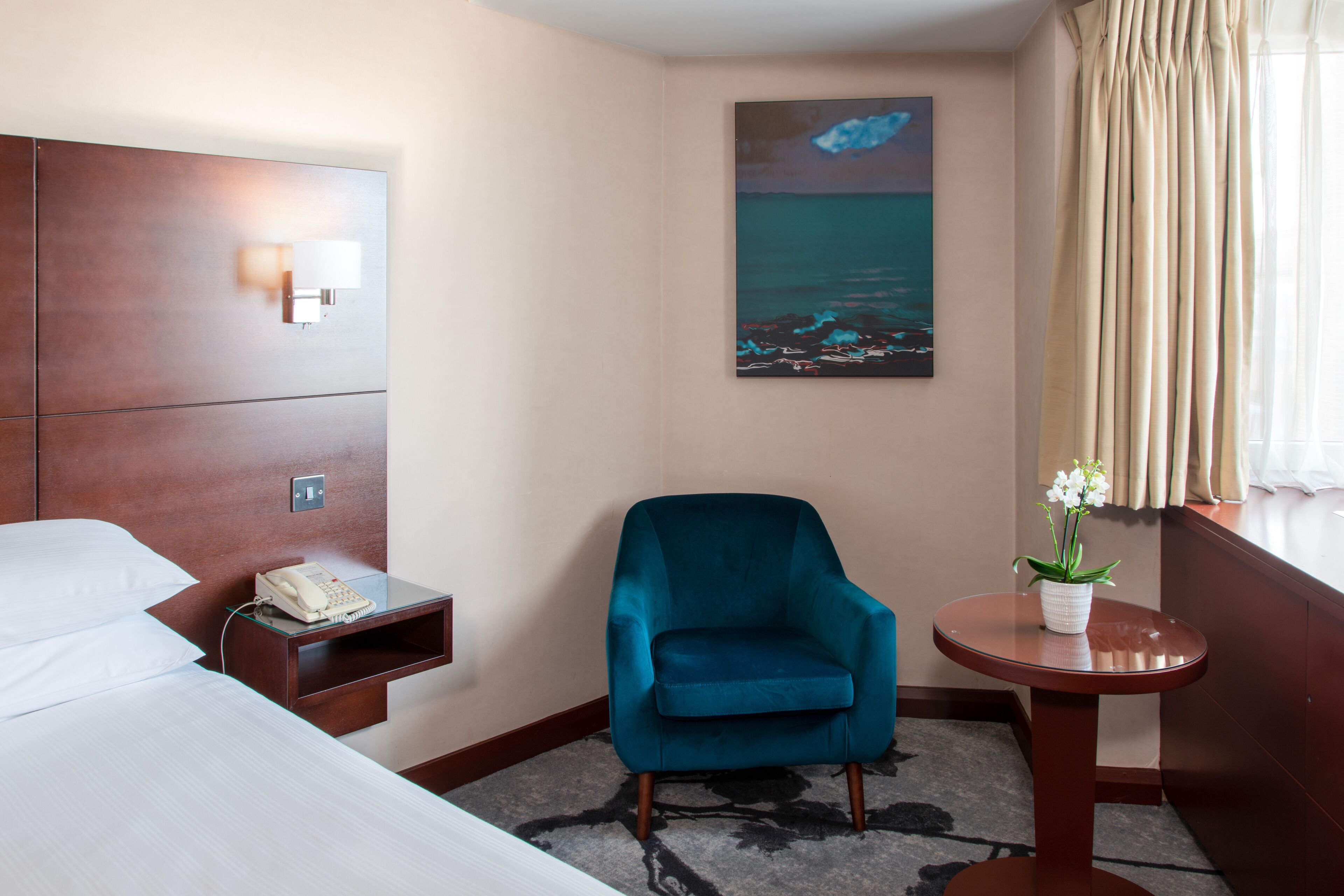 A hotel room with a bed, a desk with a phone, a teal armchair, a small table with a plant, and a painting of a night scene on the wall.