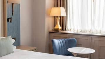 A hotel room with a bed, a blue chair, a small round table, a lamp, and a window with curtains.