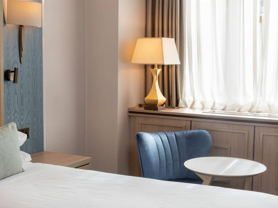A hotel room with a bed, a blue chair, a small round table, a lamp, and a window with curtains.