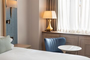 A hotel room with a bed, a blue chair, a small round table, a lamp, and a window with curtains.