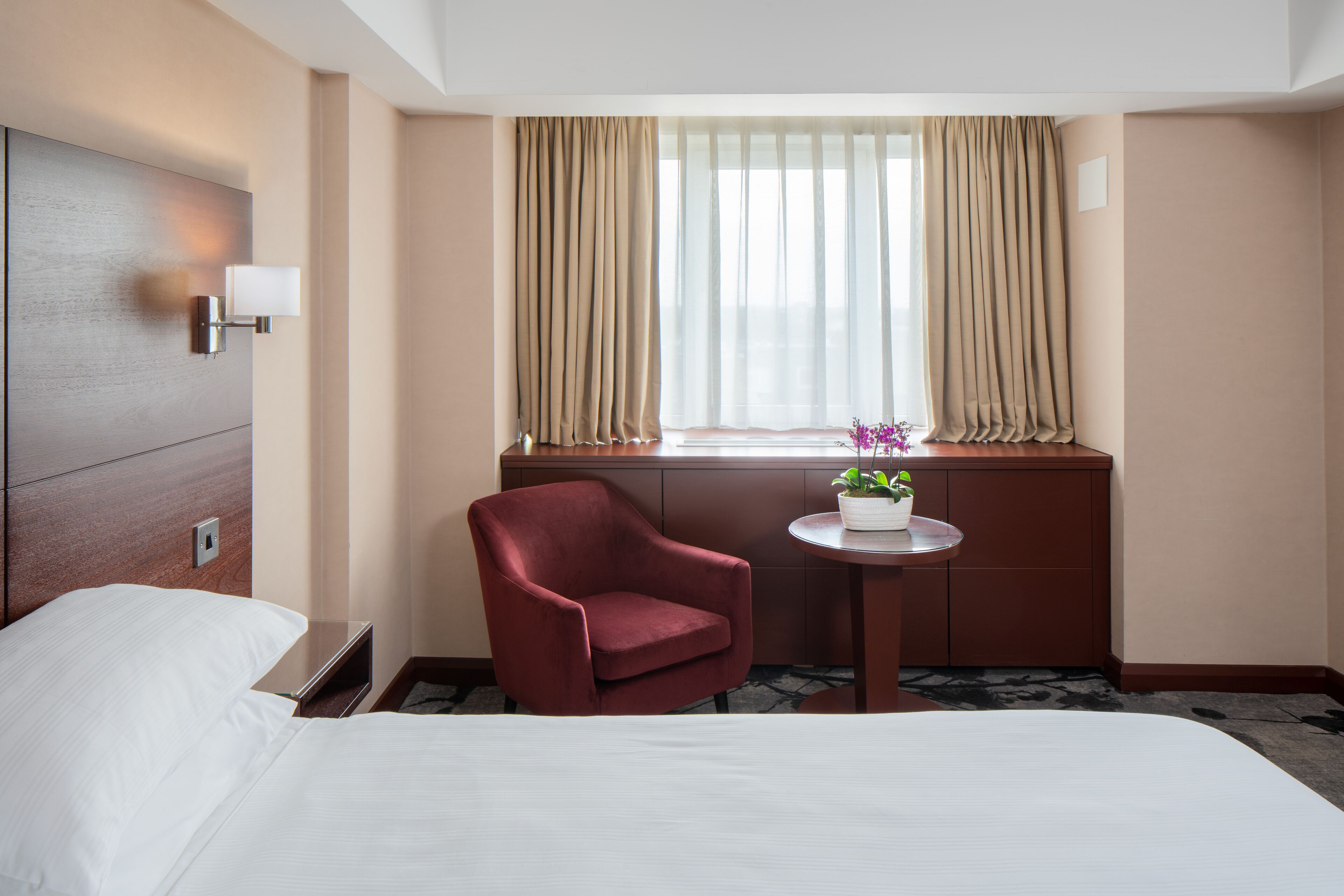 A hotel room with a bed, a red armchair, a small table with a plant, and a window with curtains.