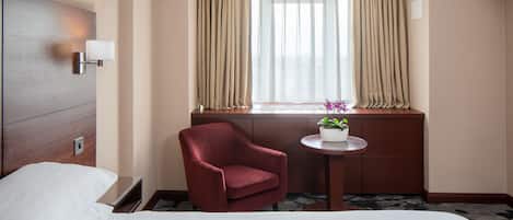 A hotel room with a bed, a red armchair, a small table with a plant, and a window with curtains.