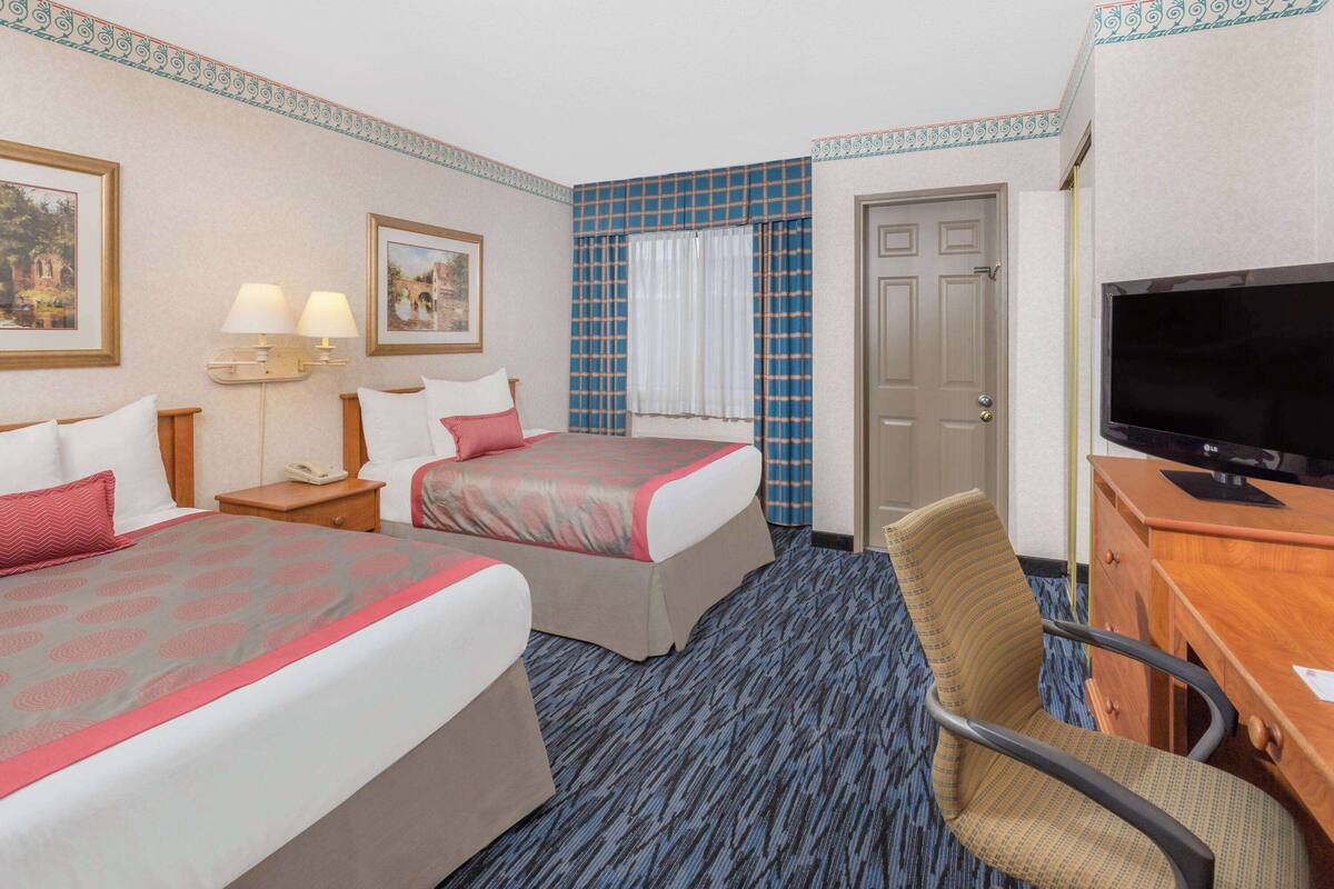 A hotel room with two beds, a desk with a TV, a chair, and a window with curtains.