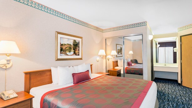 A hotel room with a large bed, bedside lamps, a nightstand, a telephone, a framed picture on the wall, and a mirror. - Ramada by Wyndham Kent Seattle Area (Kent)