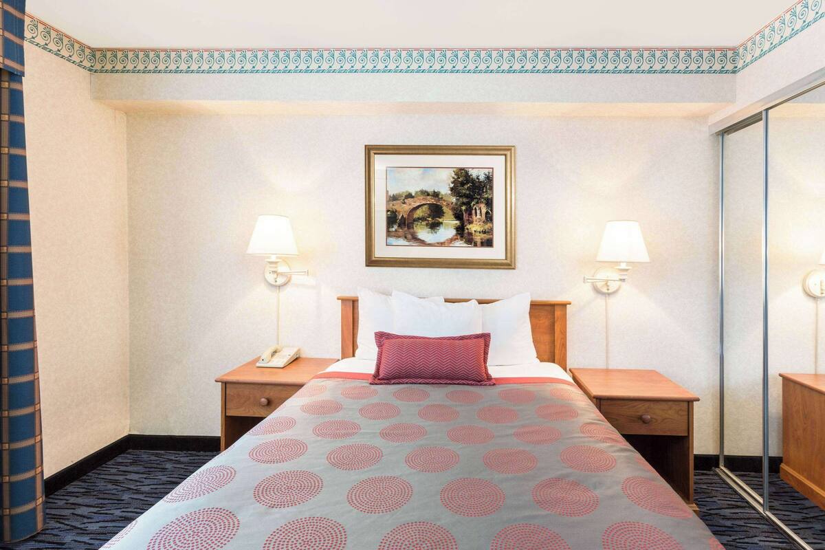 A hotel room with a bed, two bedside tables, a nightstand, a lamp, a framed picture on the wall, and a patterned bedspread.