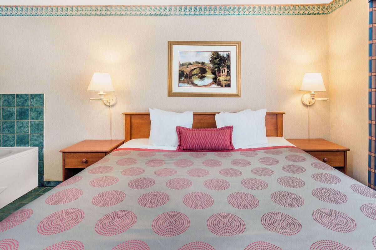 A hotel room with a bed, two nightstands, a wooden headboard, a framed picture on the wall, and a bathroom with a blue-tiled shower area.