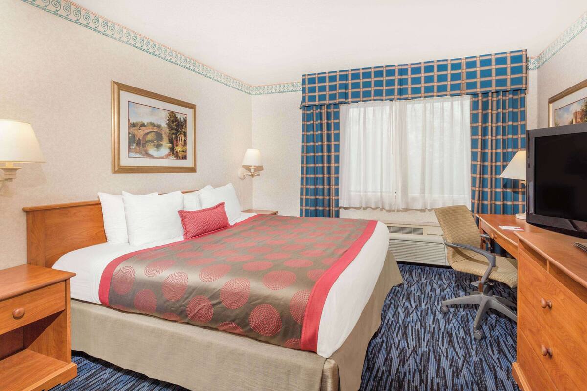 A hotel room with a large bed, a desk with a computer, a TV, and a window with curtains.