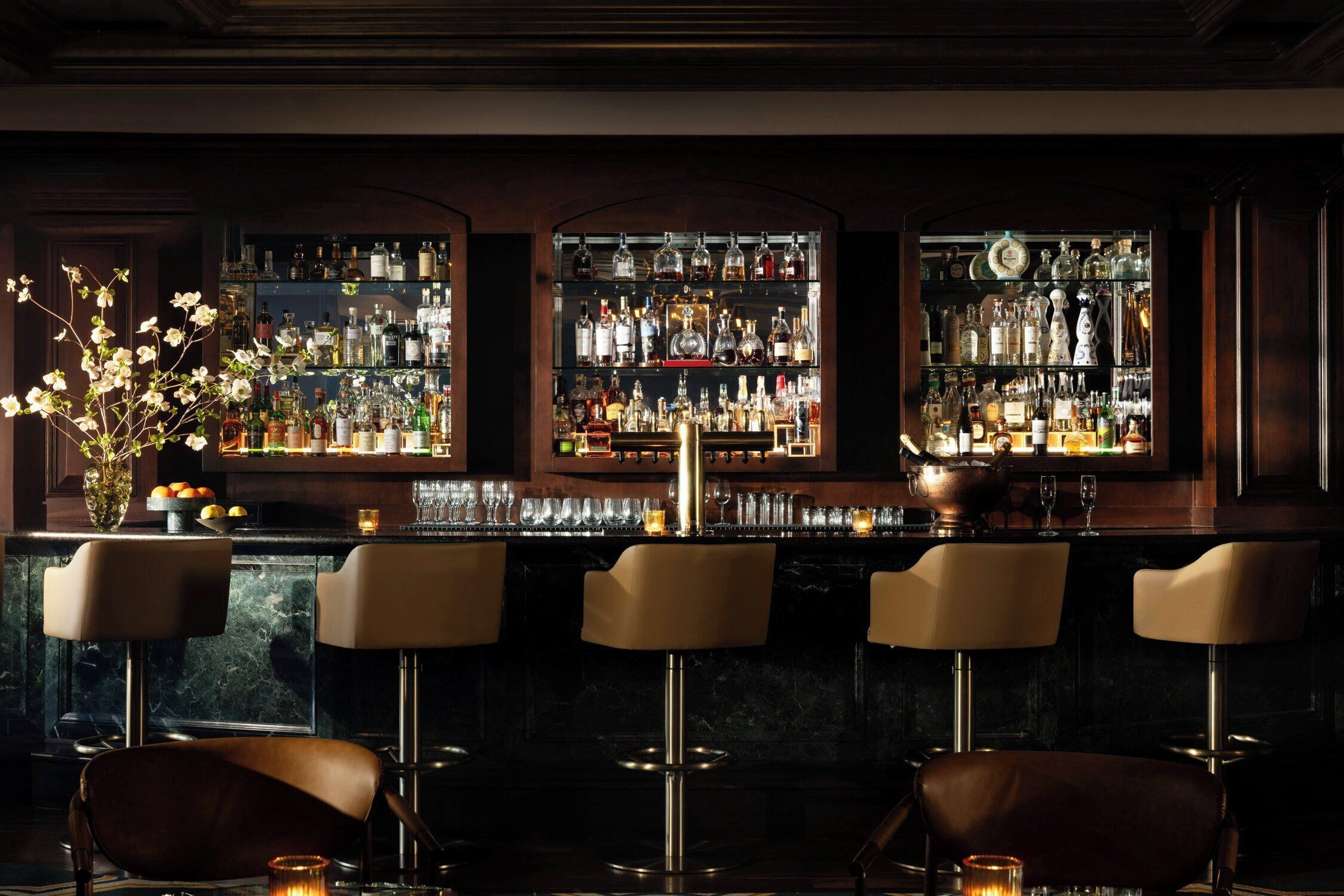 A bar area with three bar stools, a marble countertop, and shelves stocked with bottles and glasses.