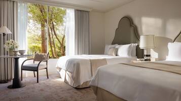 A hotel room with two beds, a desk with a lamp, a chair, and a view of trees through large windows.