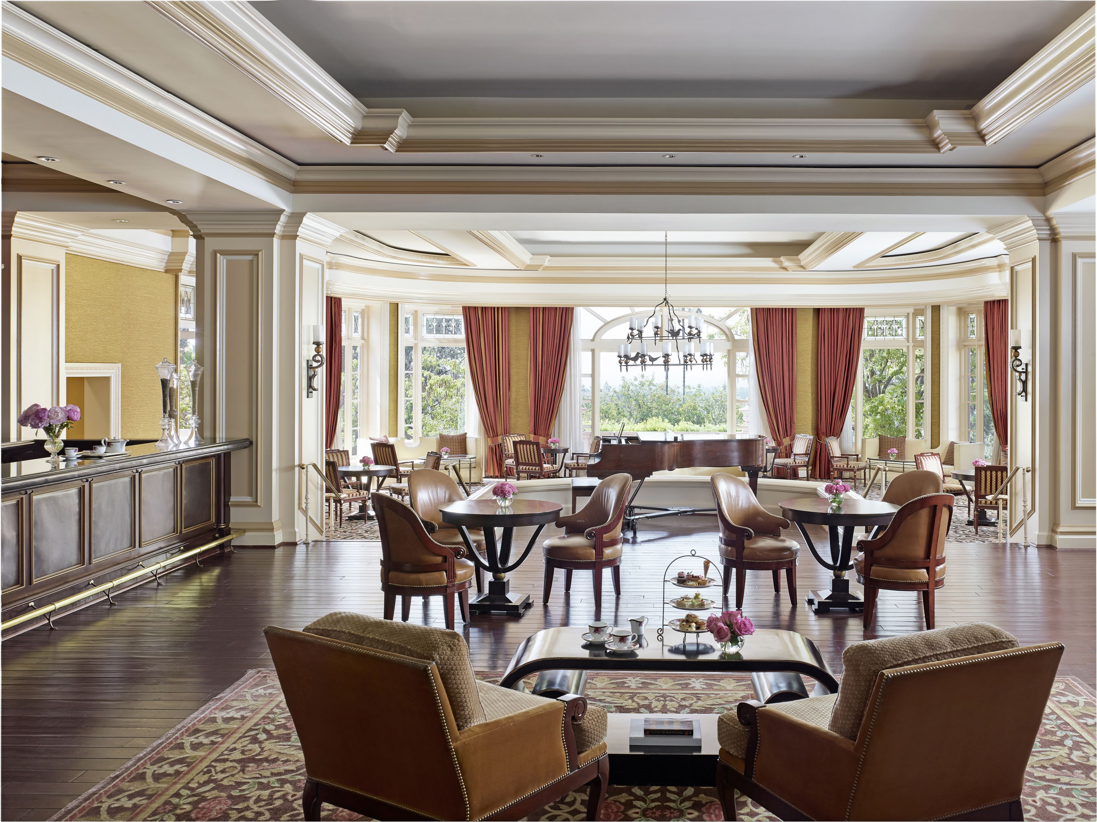 A spacious, elegantly furnished room with a grand piano, multiple seating arrangements, and large windows offering a view of greenery.