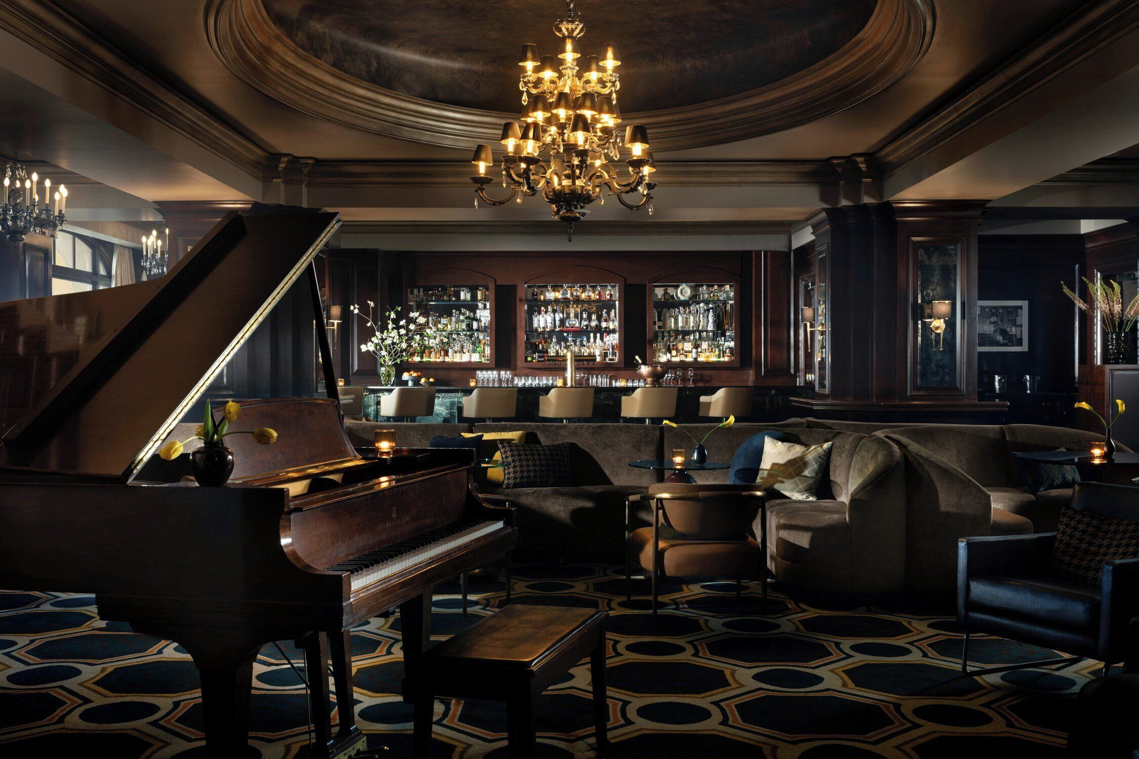 A grand piano, a bar area with shelves of bottles, a chandelier, and a patterned carpet.