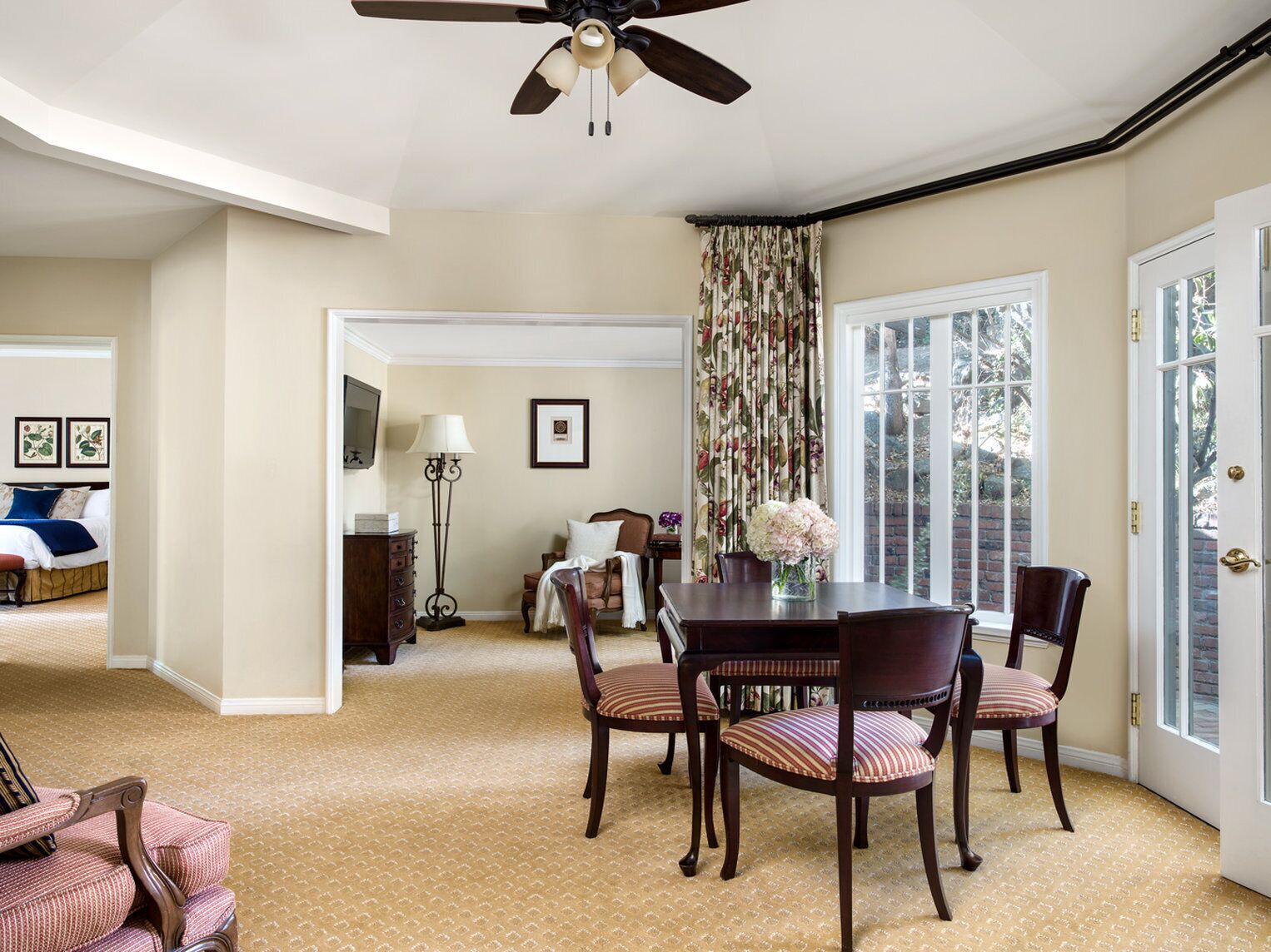 Un coin repas comprenant une table et des chaises, un ventilateur de plafond et une vue sur l’extérieur à travers une fenêtre avec des rideaux.