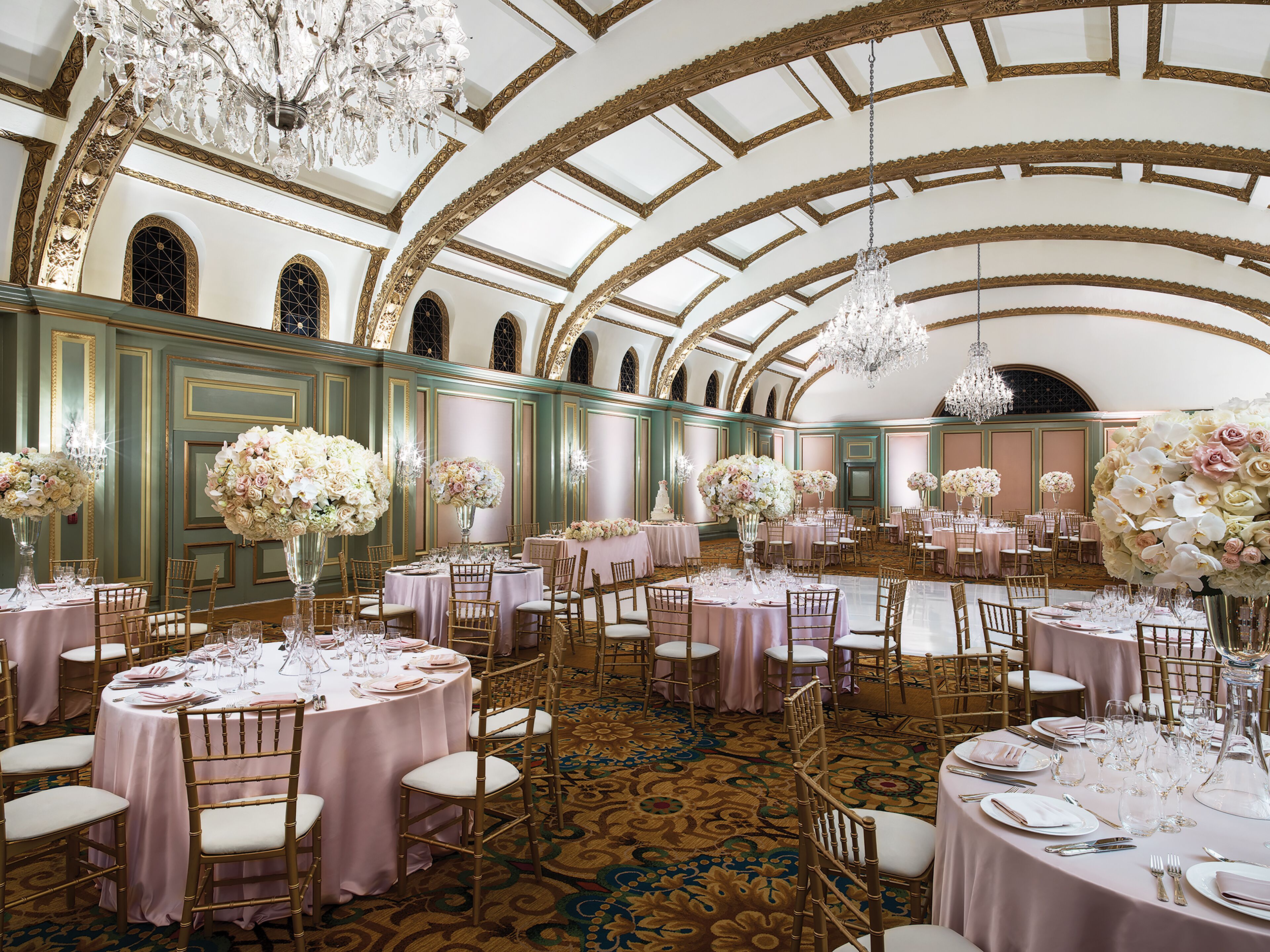 A grand ballroom with ornate chandeliers, round tables with floral centerpieces, and elegant gold chairs.