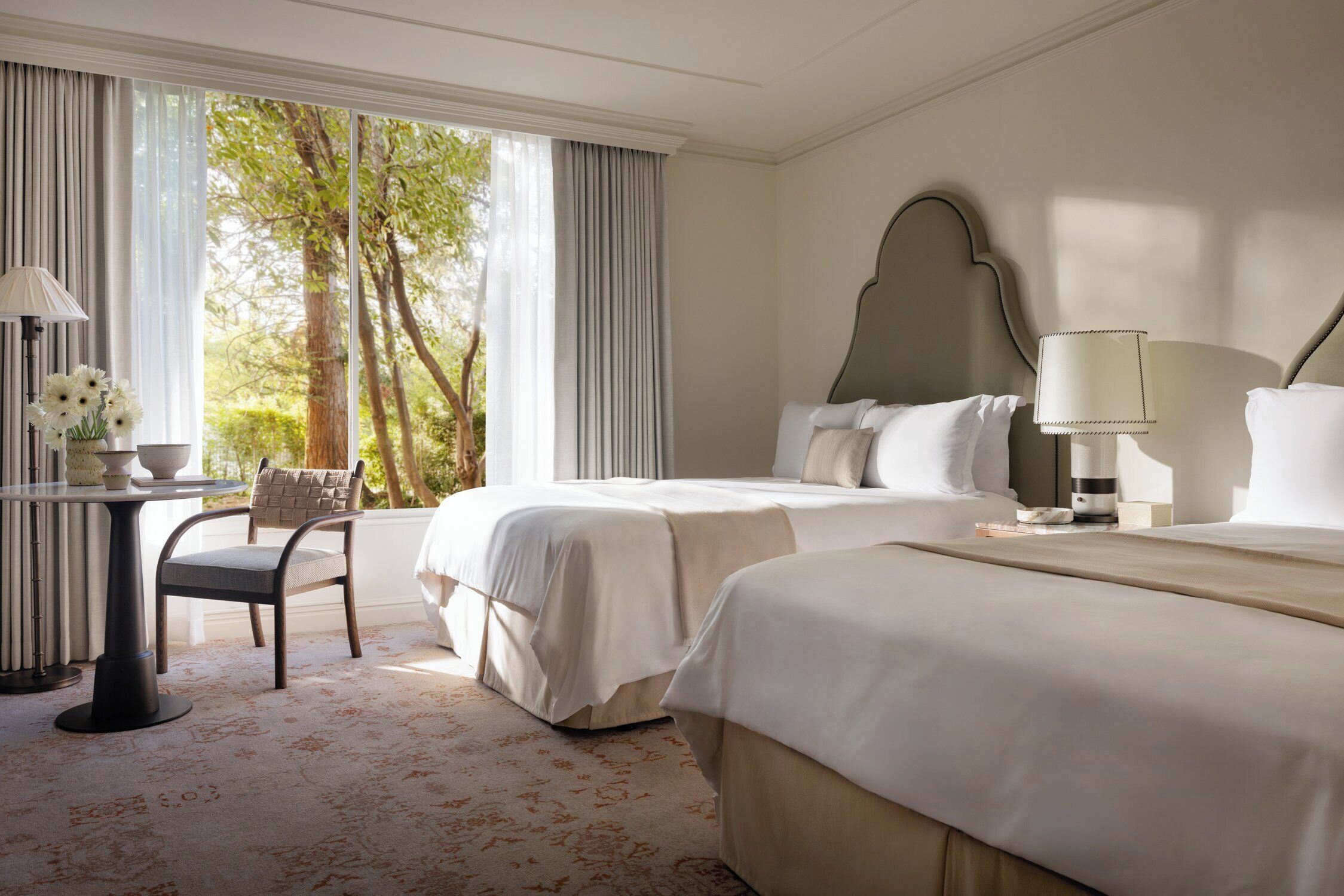 A hotel room with two beds, a desk with a lamp, a chair, and a view of trees through large windows.
