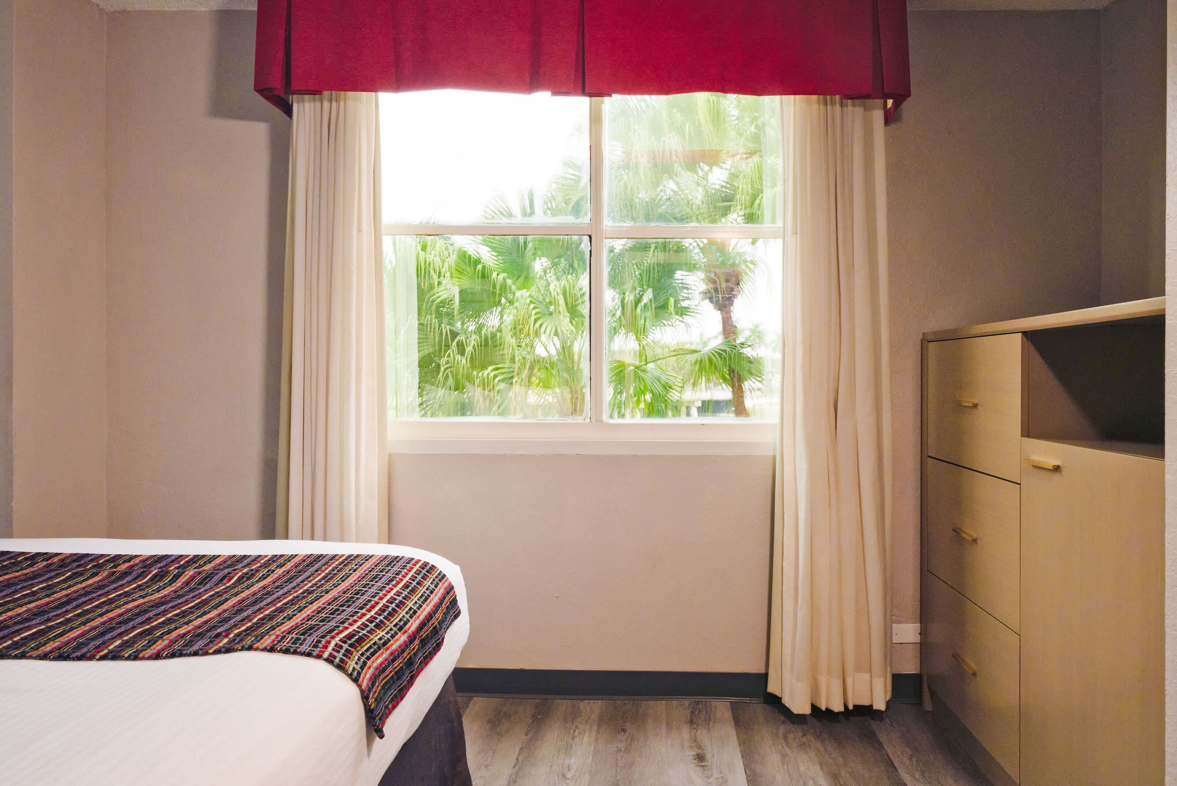 A hotel room with a bed, a window with curtains, and a wooden dresser.