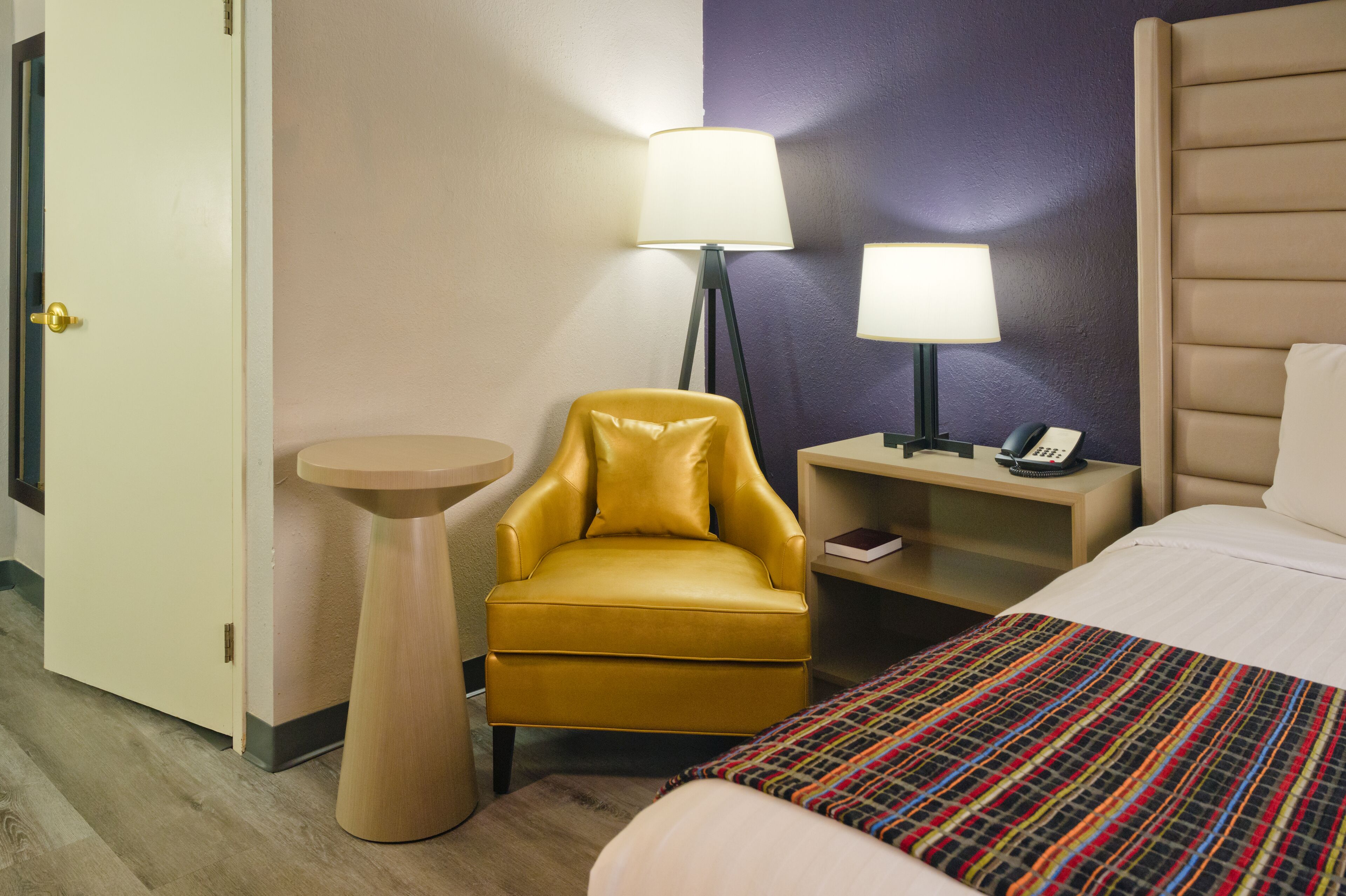 Hotellihuone, jossa on sänky, keltainen nojatuoli, yöpöytä lampun kanssa ja pieni pyöreä pöytä.