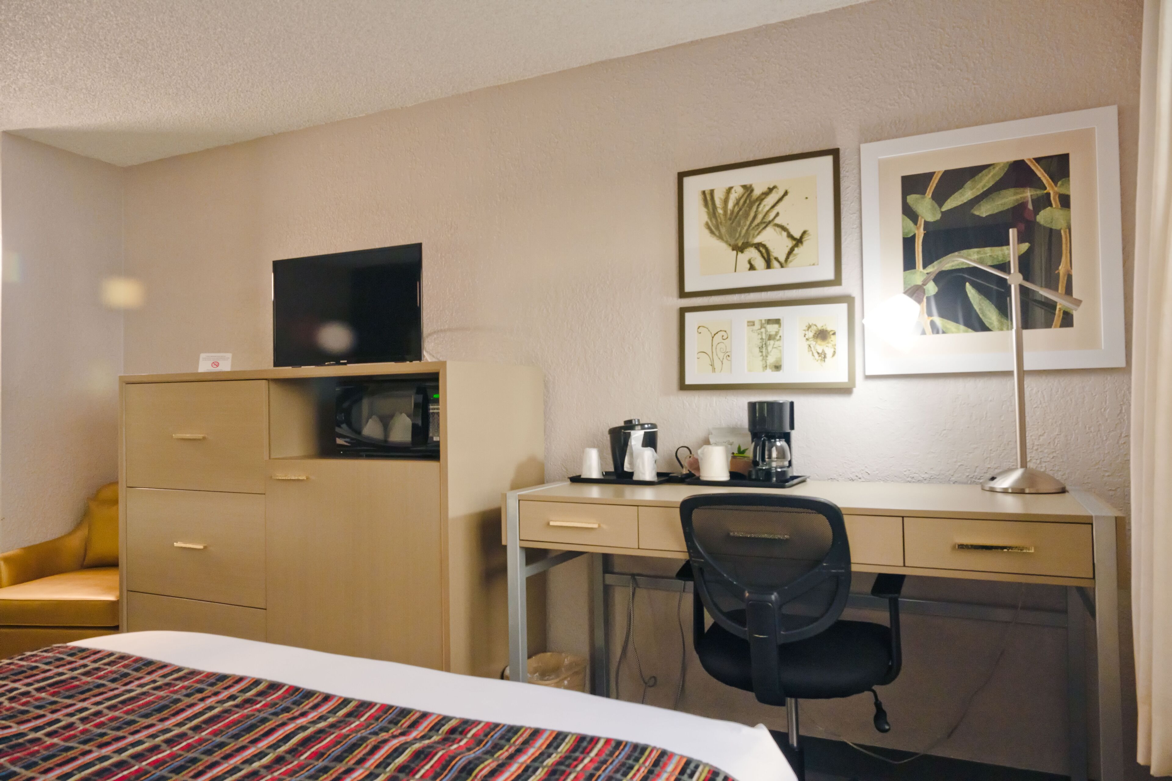 Ein Hotelzimmer mit einem Bett, einem Schreibtisch mit Stuhl, einem Fernseher auf einem Schrank und gerahmten Bildern an der Wand.
