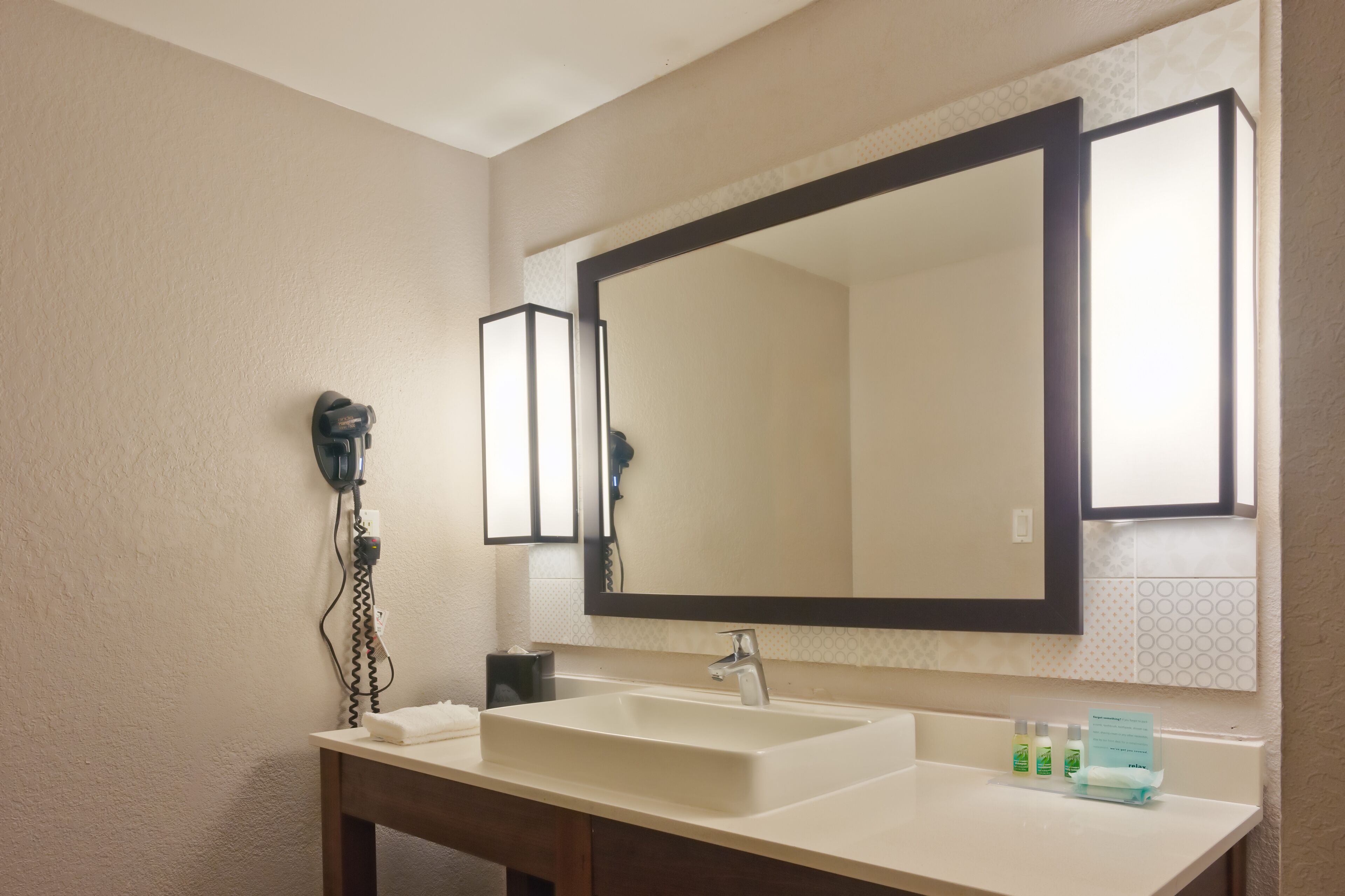 A bathroom with a large mirror, a sink, a hairdryer, and toiletries.