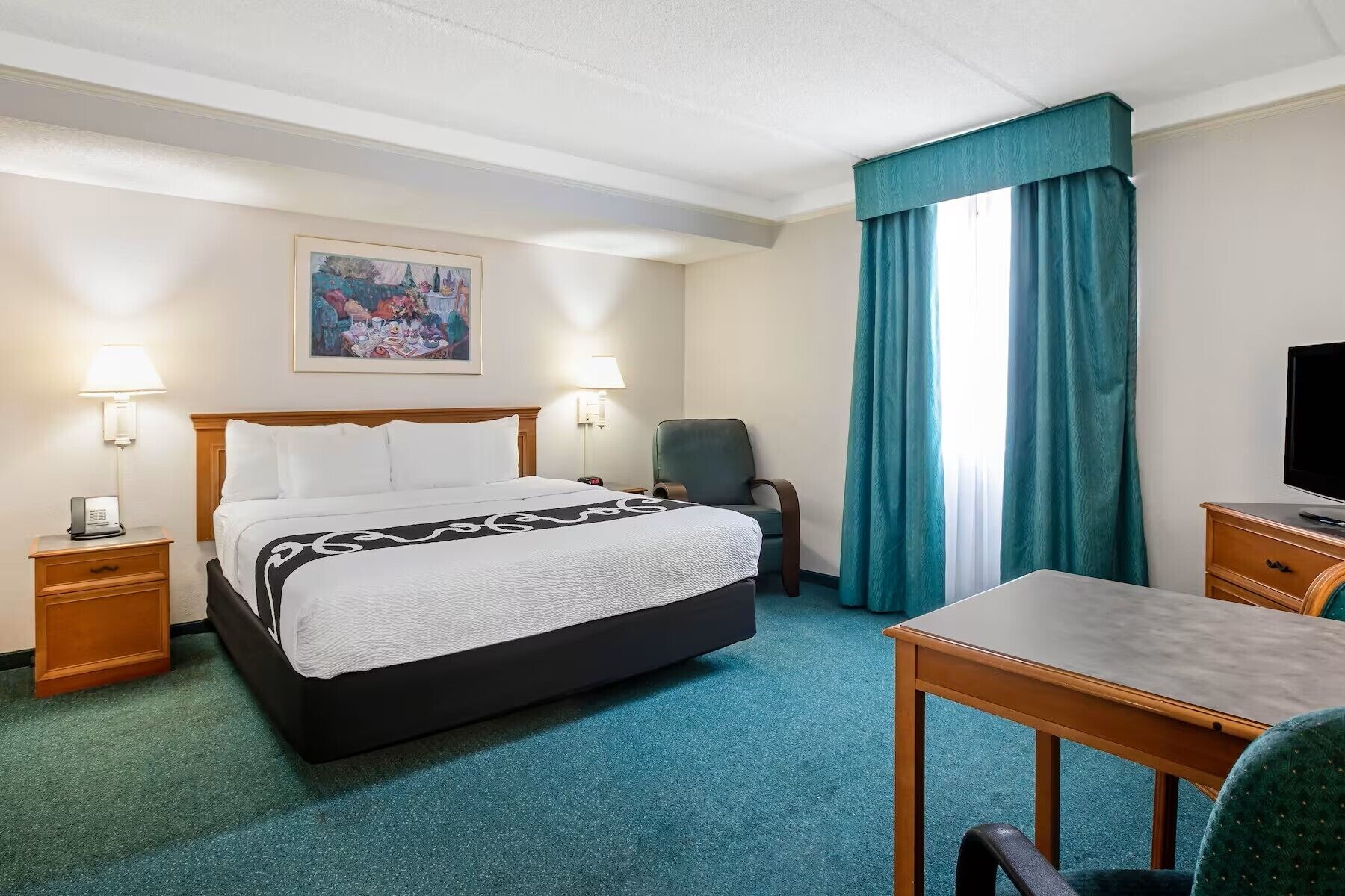 A hotel room with a bed, a desk, a chair, a television, and a painting on the wall.