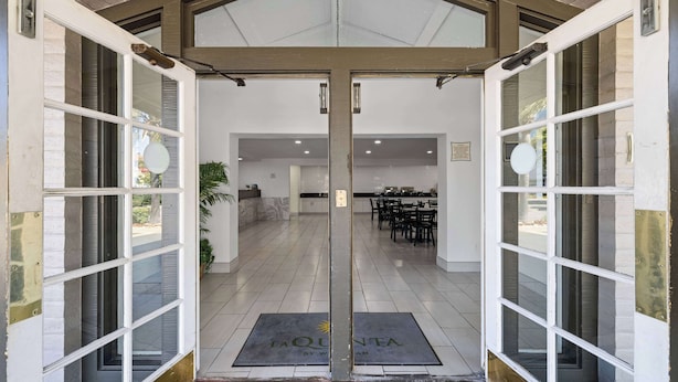 Entrada de hotel con puerta de vidrio, área de recepción con mostrador y sillas, y un cartel que dice "LA QUINTA BY WIMAN".