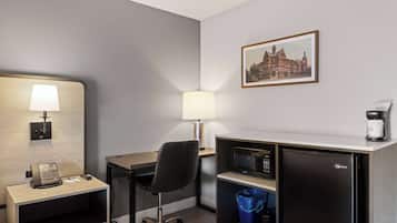 Habitación de hotel con escritorio, silla, lámpara, microondas, refrigerador y un cubo de basura.