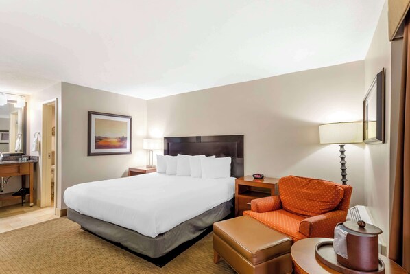 A hotel room with a large bed, a desk with a lamp, an orange chair, and a small table with a teapot.