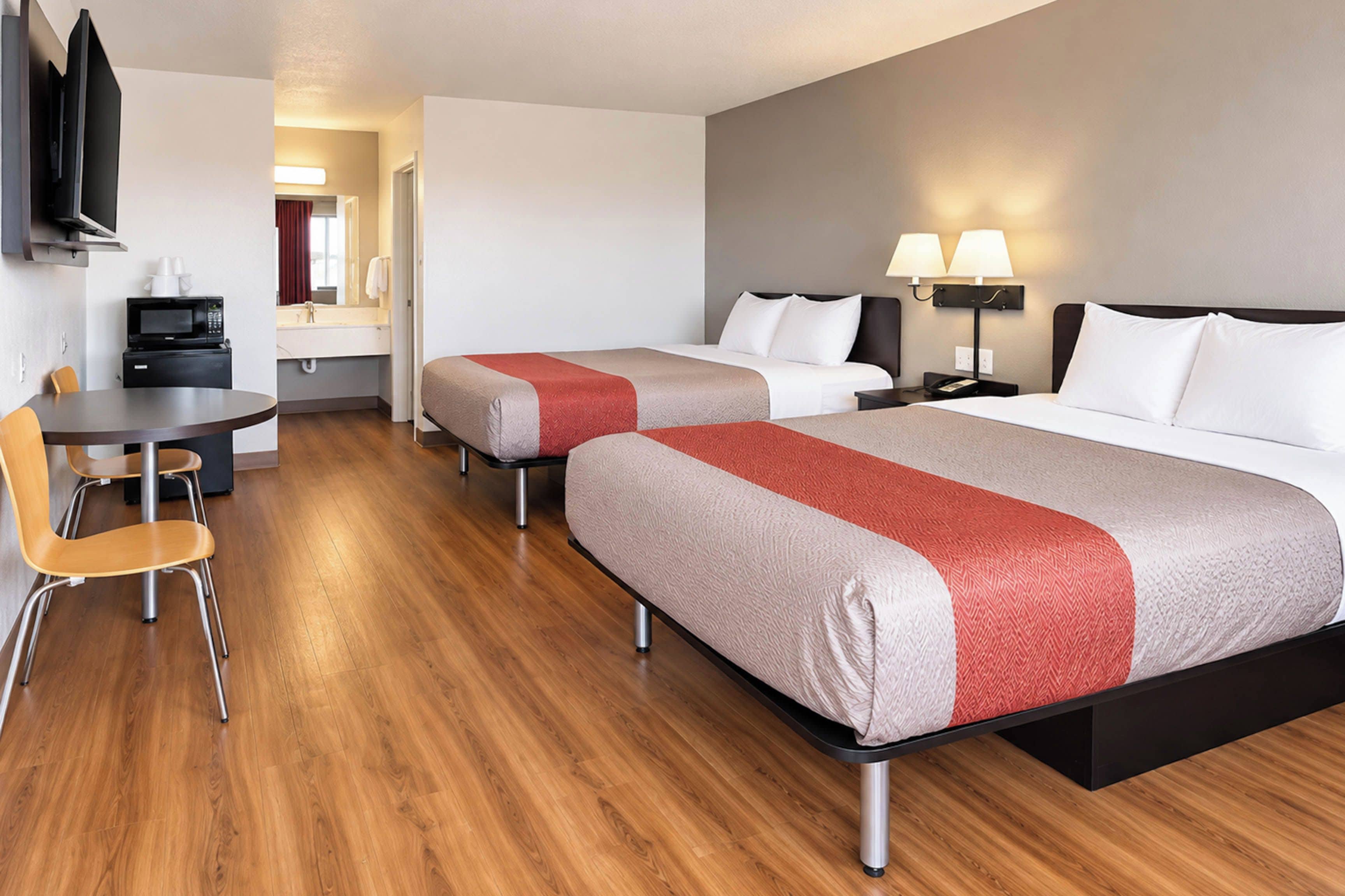 A hotel room with two beds, a desk, a chair, a TV, and a bathroom visible in the background.