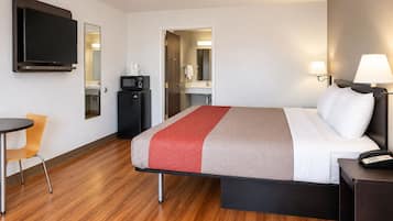 A hotel room with a bed, bedside table, lamp, TV, microwave, and a small desk with chair.