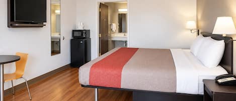 A hotel room with a bed, bedside table, lamp, TV, microwave, and a small desk with chair.