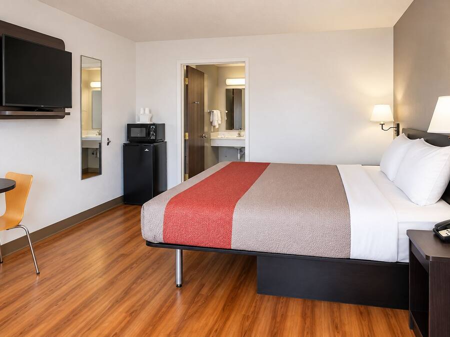 A hotel room with a bed, bedside table, lamp, TV, microwave, and a small desk with chair.