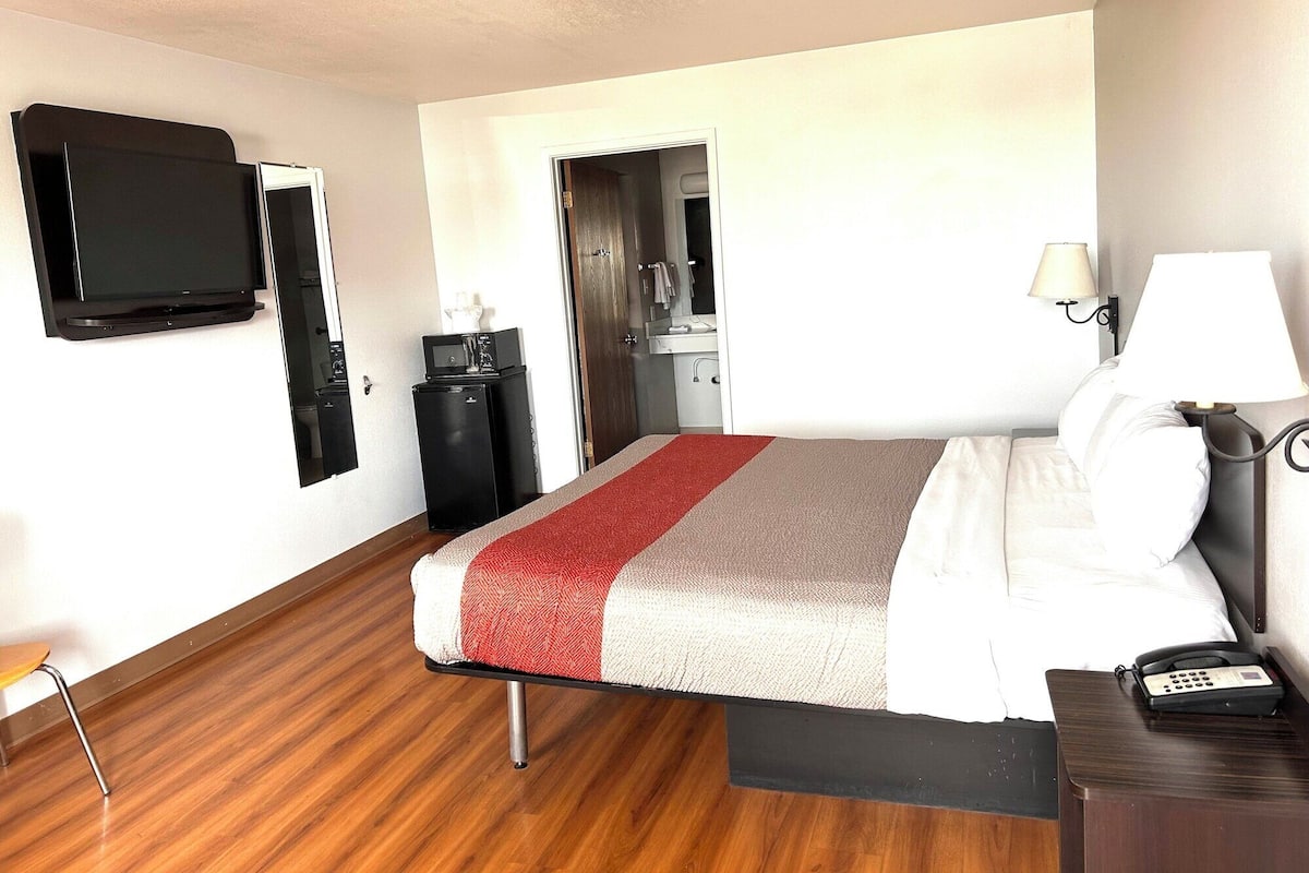 A hotel room with a bed, a desk with a telephone, a television mounted on the wall, and a small refrigerator.