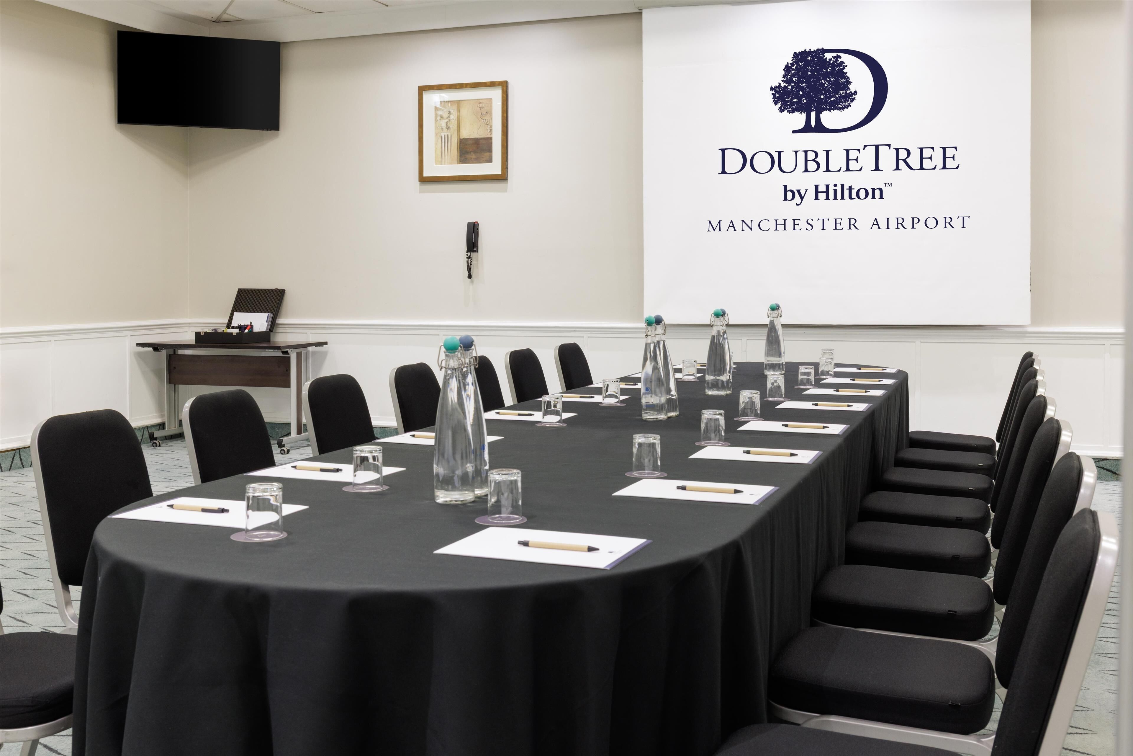 A conference room with a long table, chairs, water bottles, and a projector screen displaying the DoubleTree by Hilton logo at Manchester Airport.