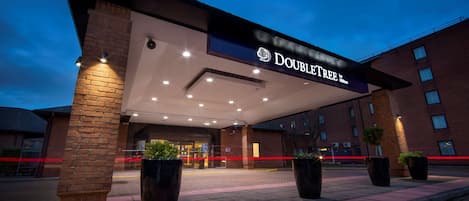 Een hotelentree met een opvallend bord, grote plantenbakken en een gevel van baksteen.