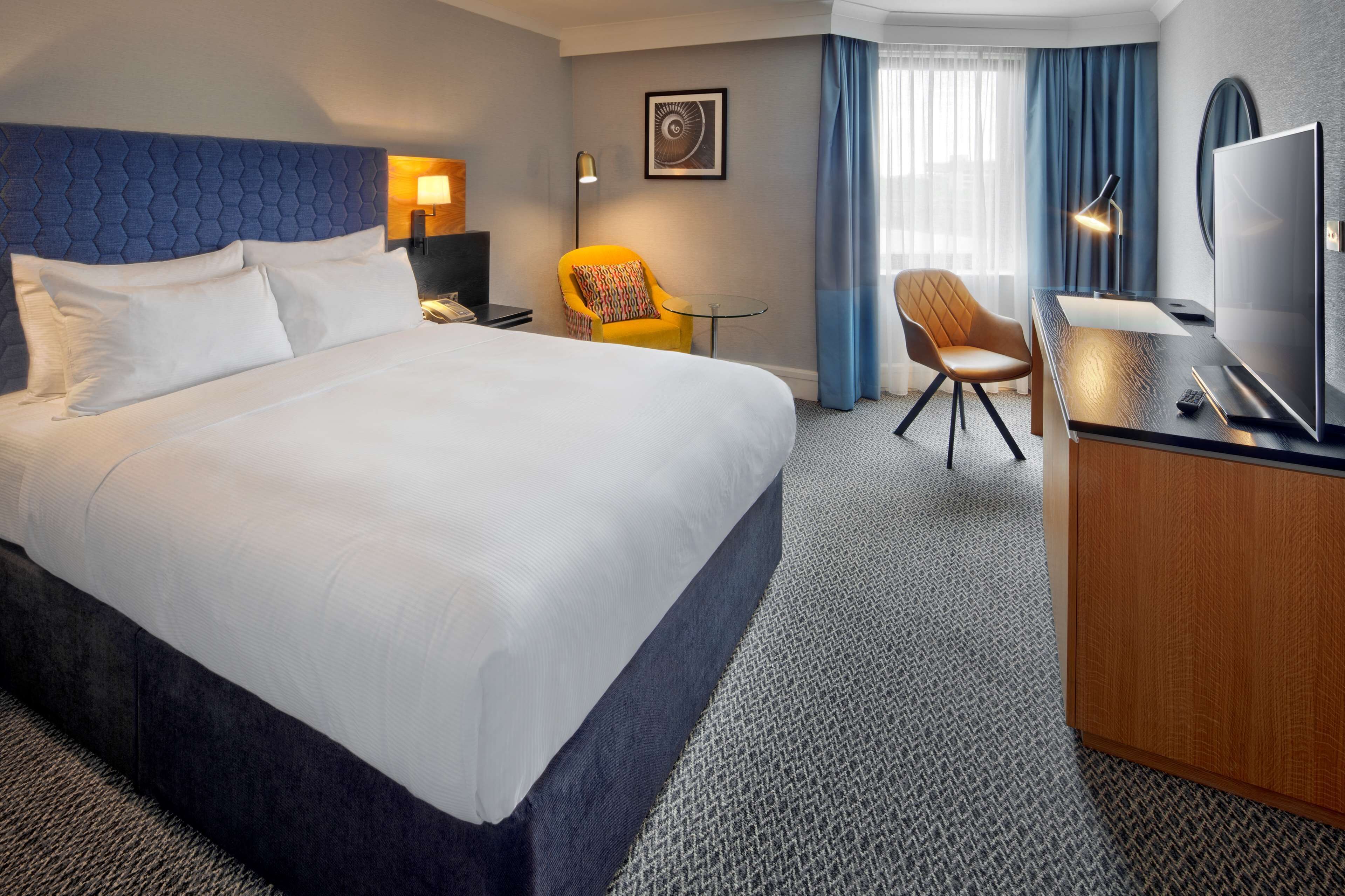 Ein Hotelzimmer mit einem großen Bett, einem Schreibtisch mit Fernseher, einem Sessel und einer Lampe.