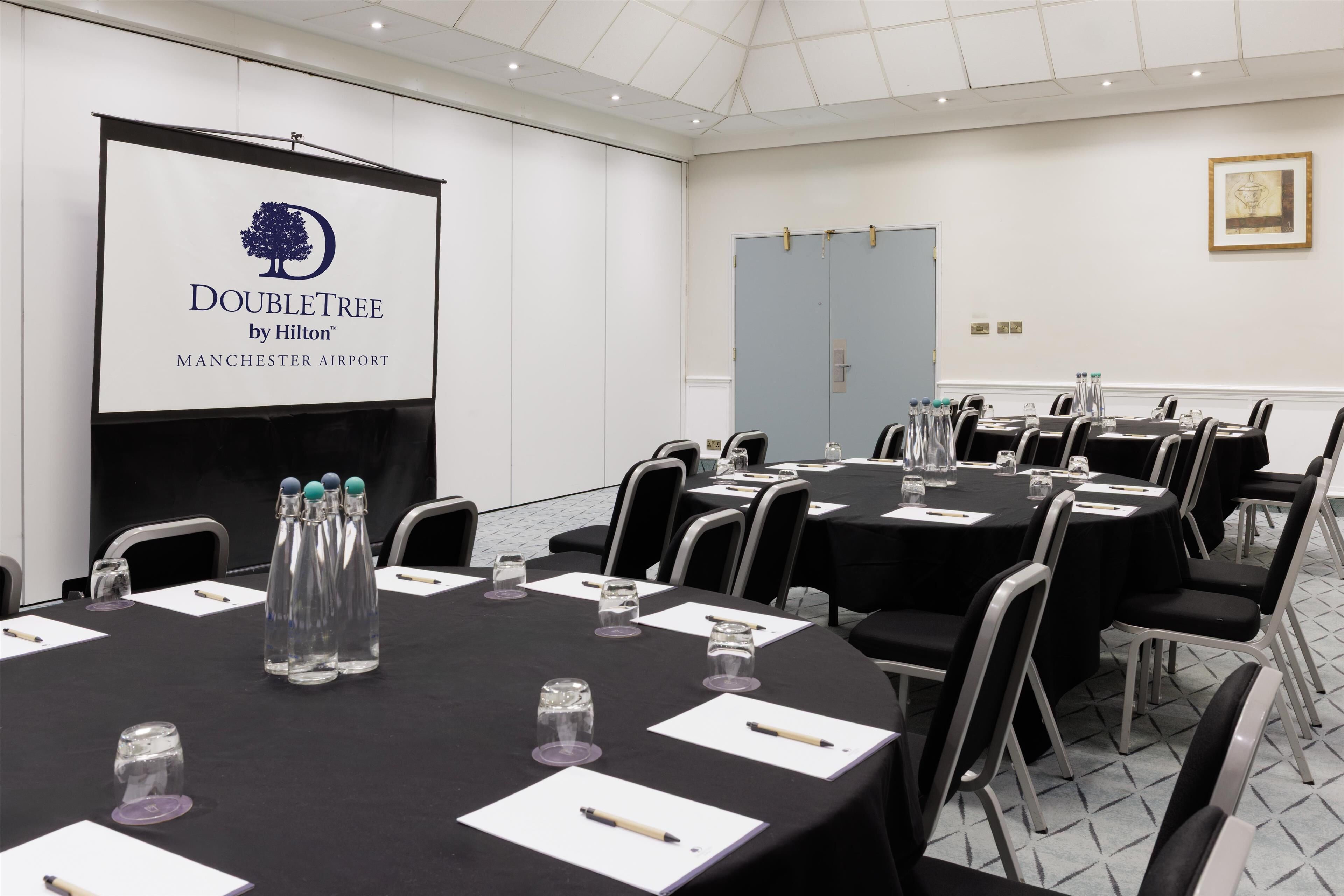 Een vergaderzaal met aanwezige tafels, een scherm waarop het logo van DoubleTree by Hilton wordt getoond, en een deur op de achtergrond.