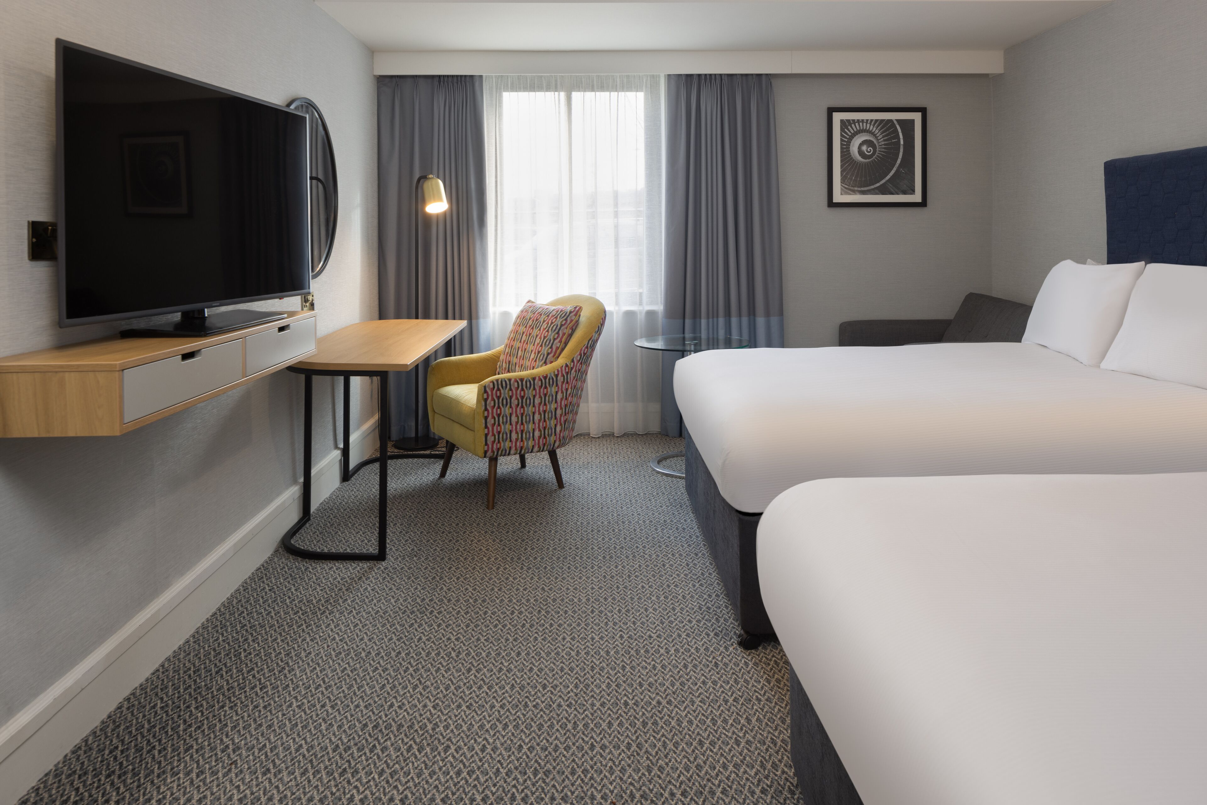 Een hotelkamer met een flatscreen-tv, een klein bureau, een stoel, een bed en een raam met gordijnen.