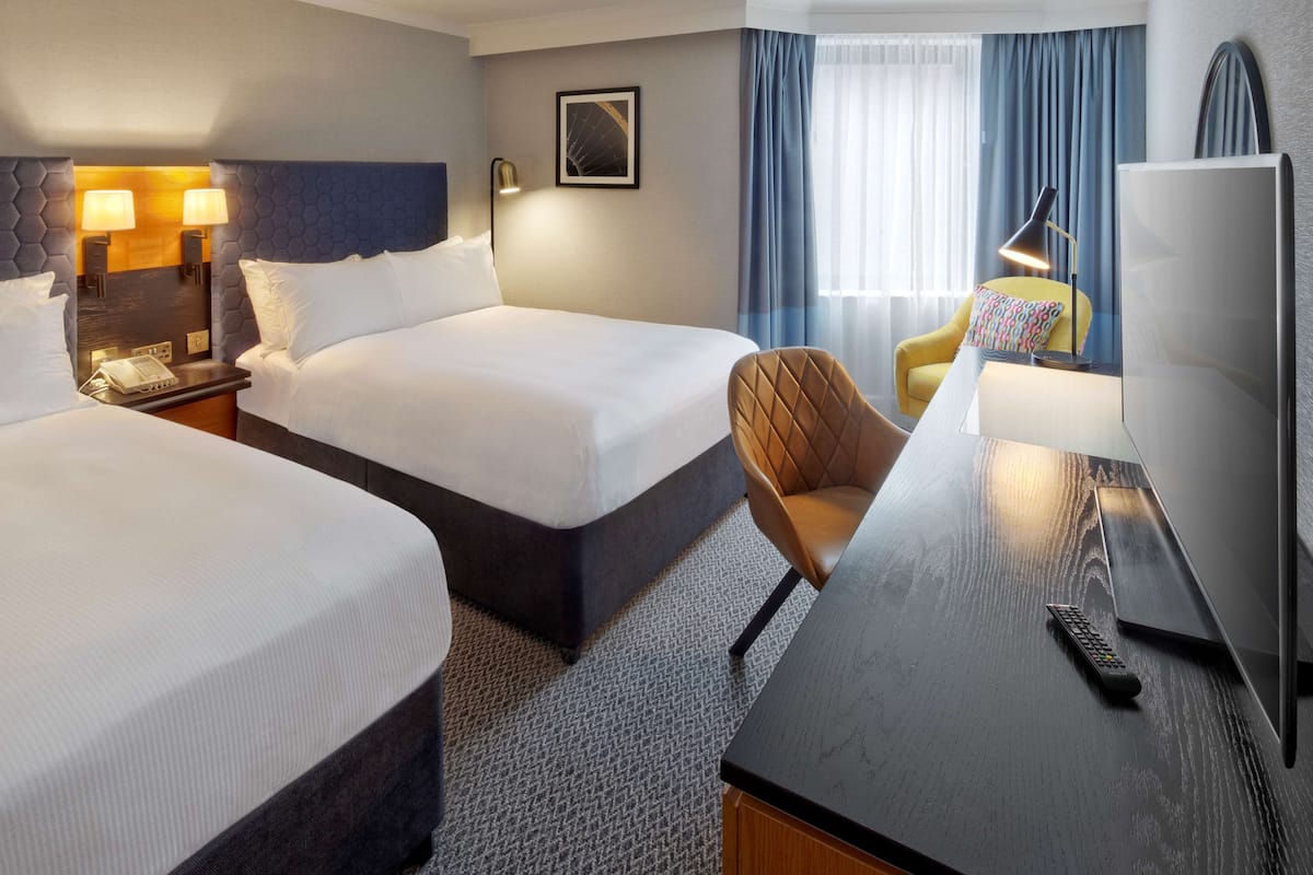 A hotel room with two beds, a desk with a lamp, a chair, and a TV.