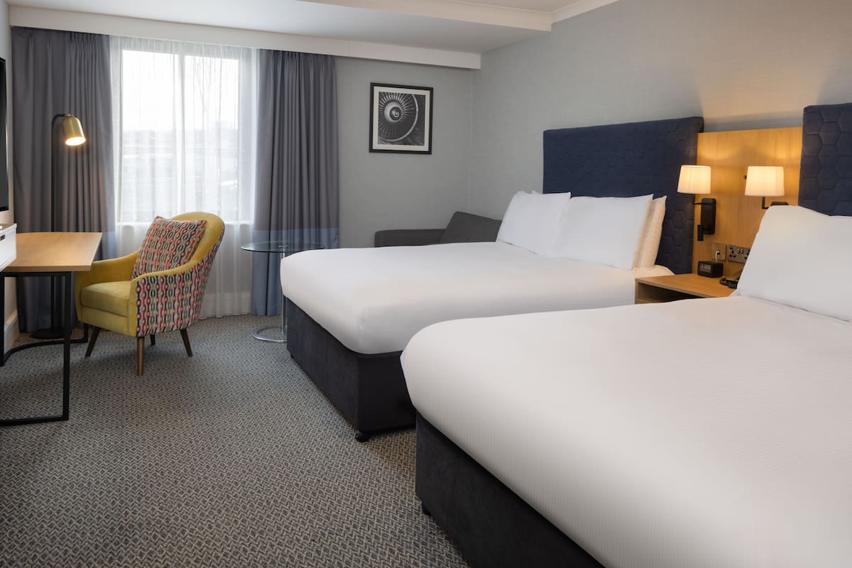 A hotel room with two beds, a desk, a chair, and a large window with curtains.