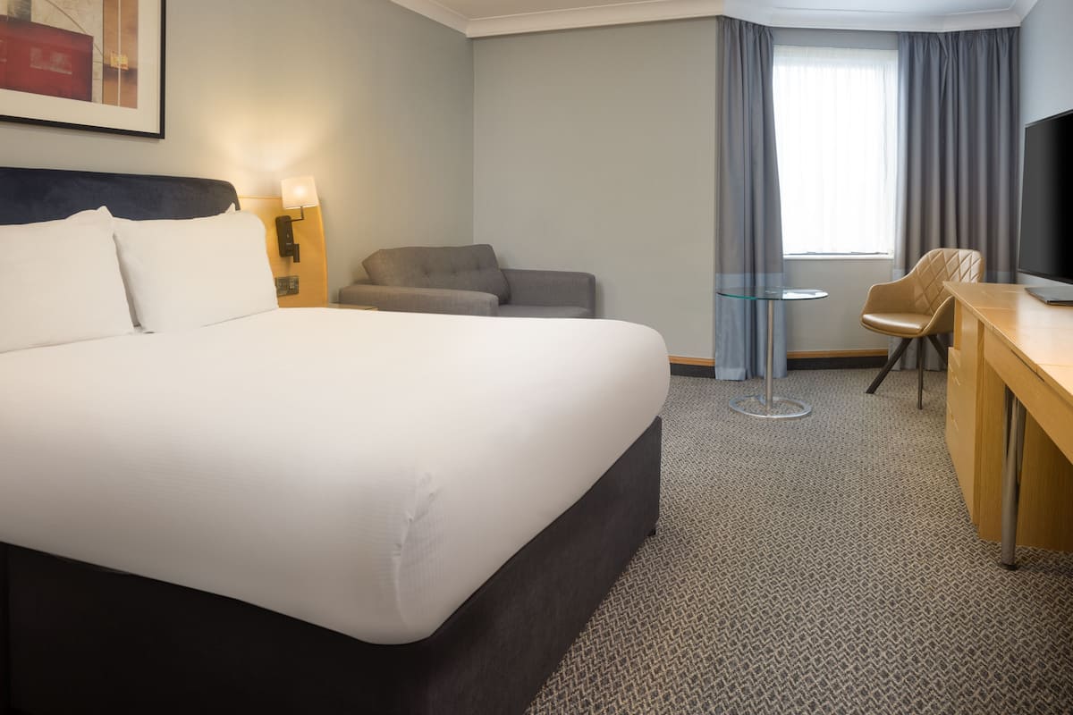 A hotel room with a large bed, a desk with a chair, a small table, a TV, and a window with curtains.