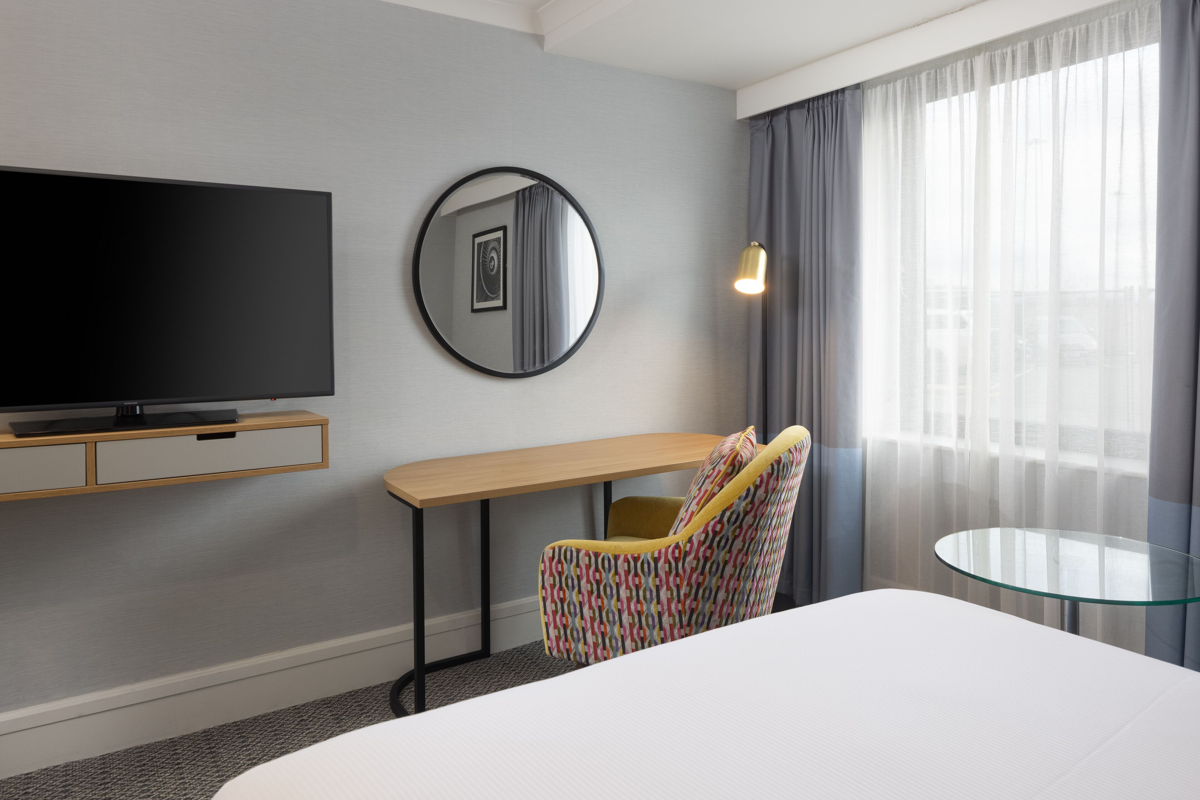 A hotel room with a bed, a TV, a round mirror, a desk, a chair, and a glass table.