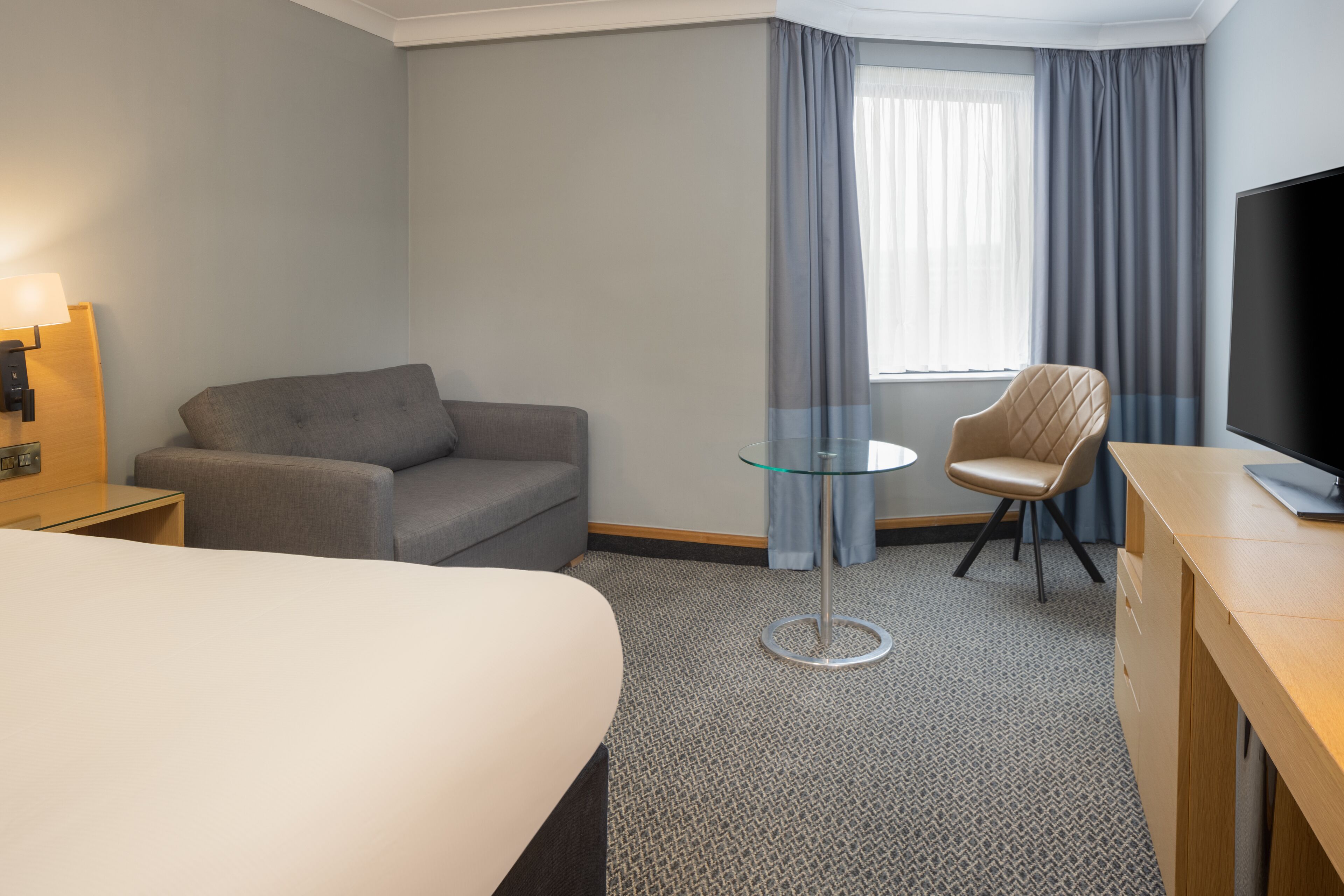 A hotel room with a bed, a grey sofa, a small glass table, a chair, a TV, and a window with curtains.