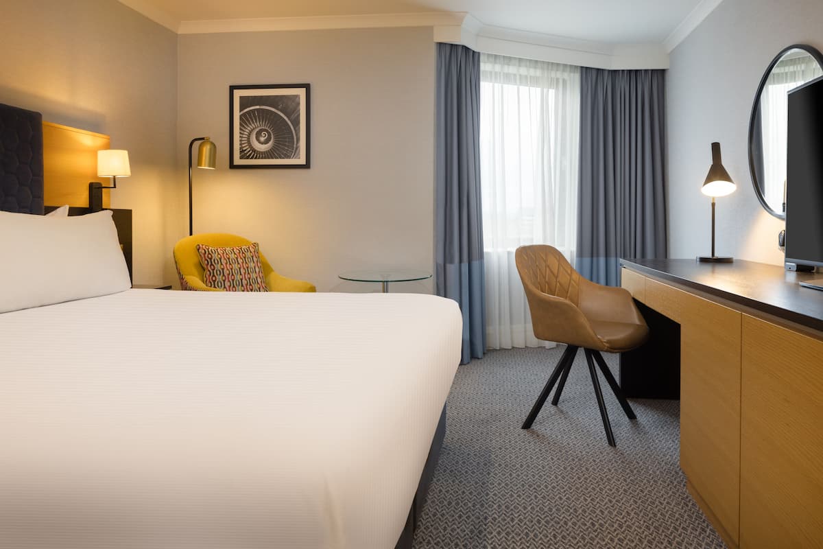 A hotel room with a large bed, a desk, a chair, a small table, a lamp, a framed picture, and a window with curtains.