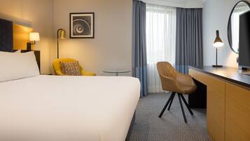 Una habitación de hotel con una cama grande, un escritorio, una silla, una mesita, una lámpara, un cuadro enmarcado y una ventana con cortinas.