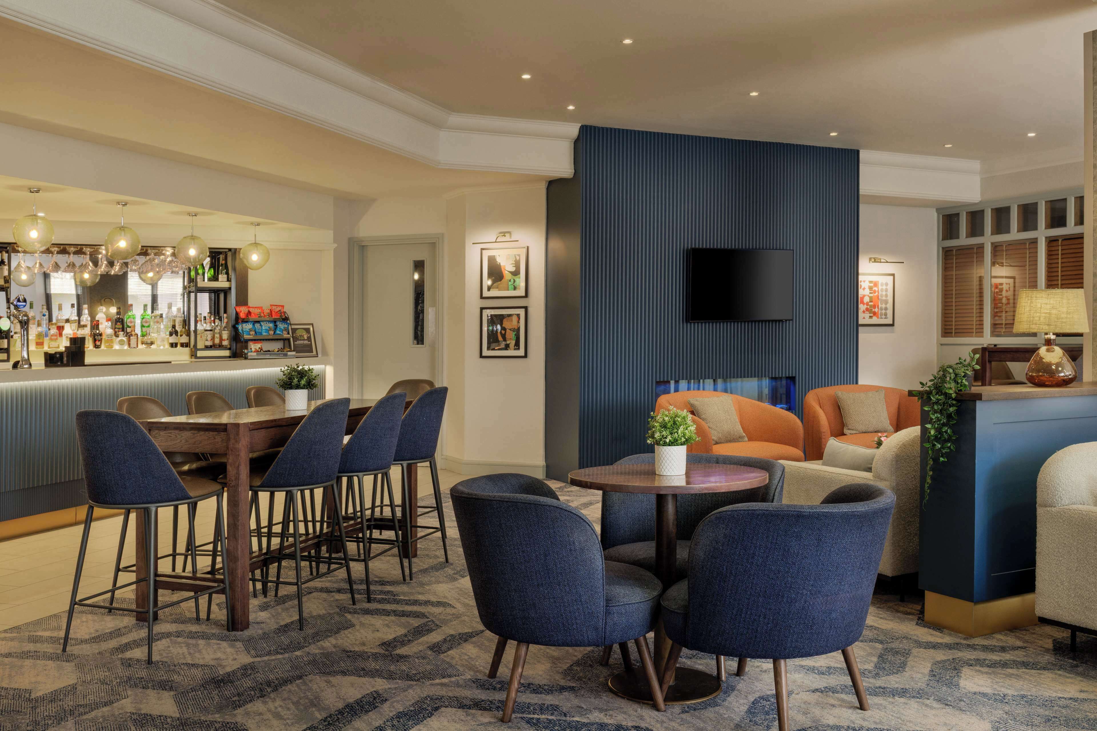 Eine Hotelhalle mit Barbereich, Sitzgelegenheiten mit blauen Stühlen, einem runden Tisch mit Pflanze und einer großen, blauen Akzentwand.